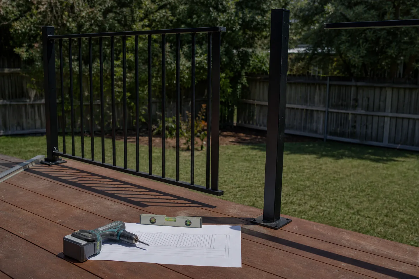 A printed balustrade drawing weighted down on a timber deck beside a half-installed black aluminium flat-top bar balustrade