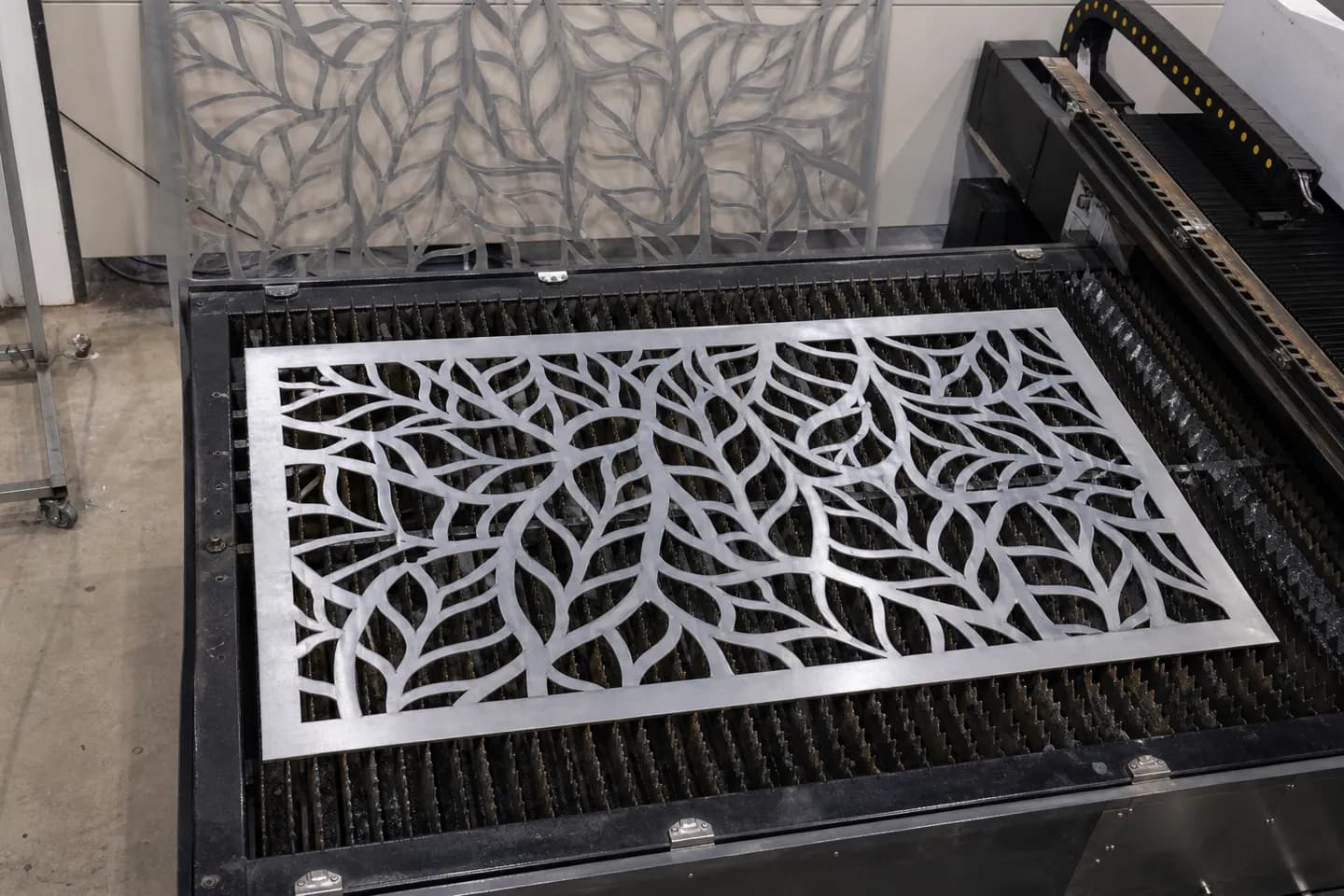 A freshly laser-cut aluminium screen panel with a leaf pattern of cut-outs lying on the slats of a flatbed laser