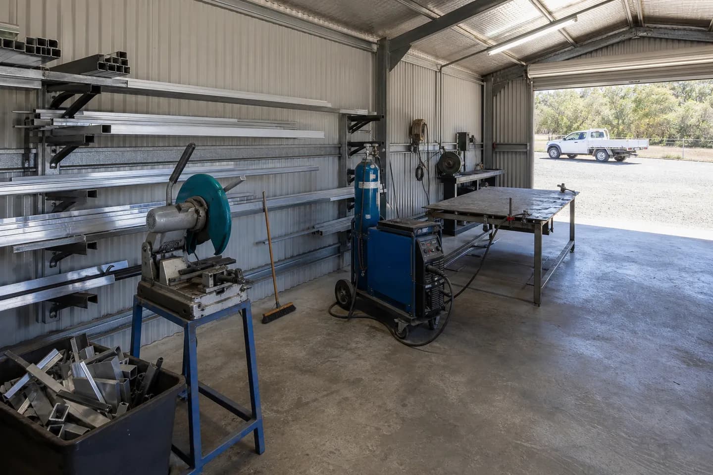 Inside a small Australian fabrication workshop with a cold saw, a welding machine on a trolley and racks of aluminium tube
