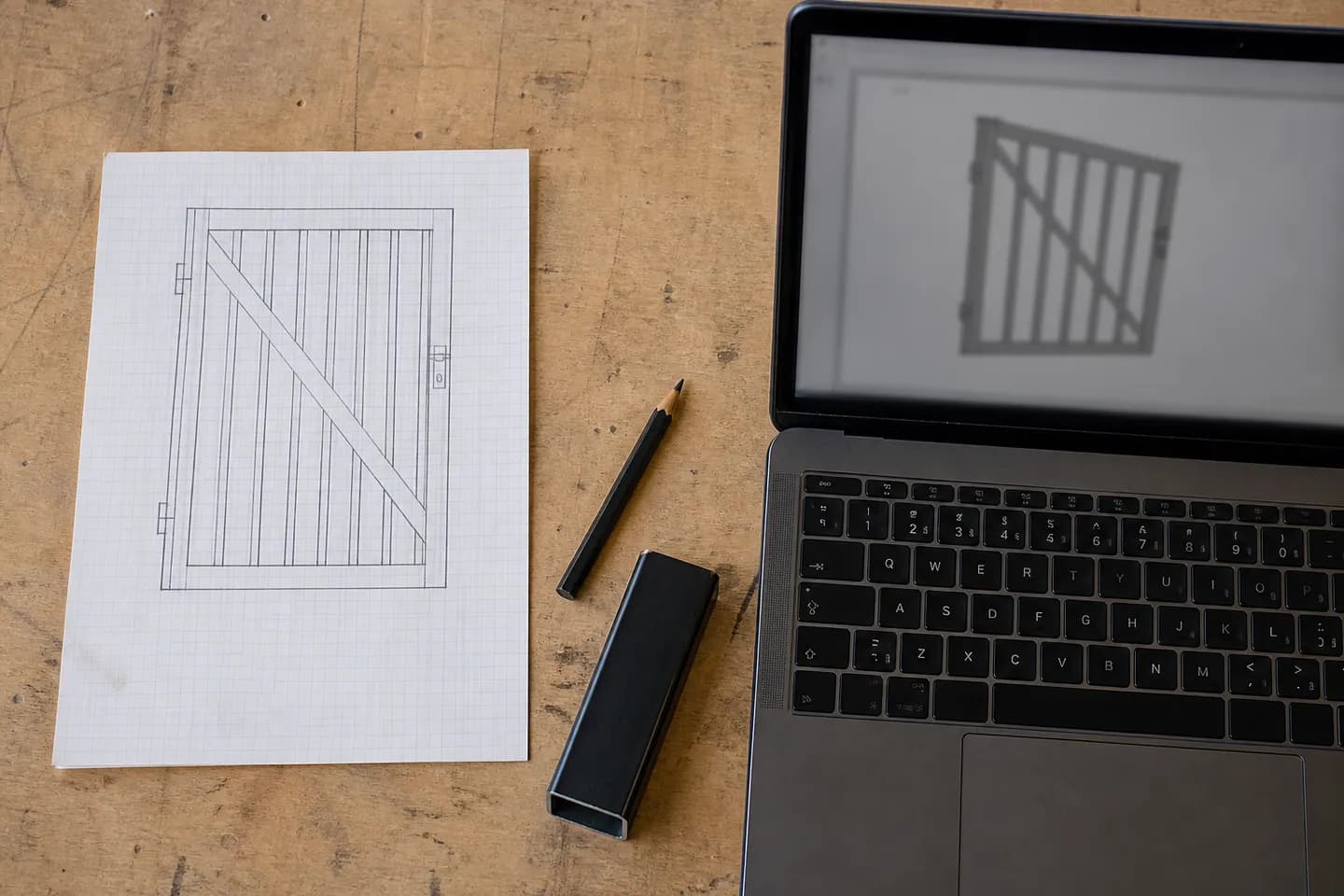 A pencil sketch of a battened gate on graph paper next to an open laptop showing a blurred 3D gate model on a workshop desk