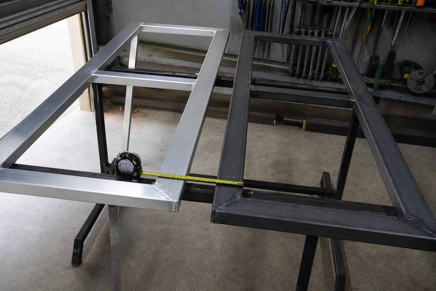 Two welded gate frames side by side on workshop trestles, one bright silver aluminium and one dark grey galvanised
