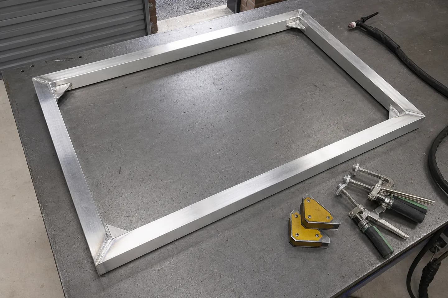 A welded aluminium square tube frame with mitred corners and small triangular gussets lying on a workshop bench