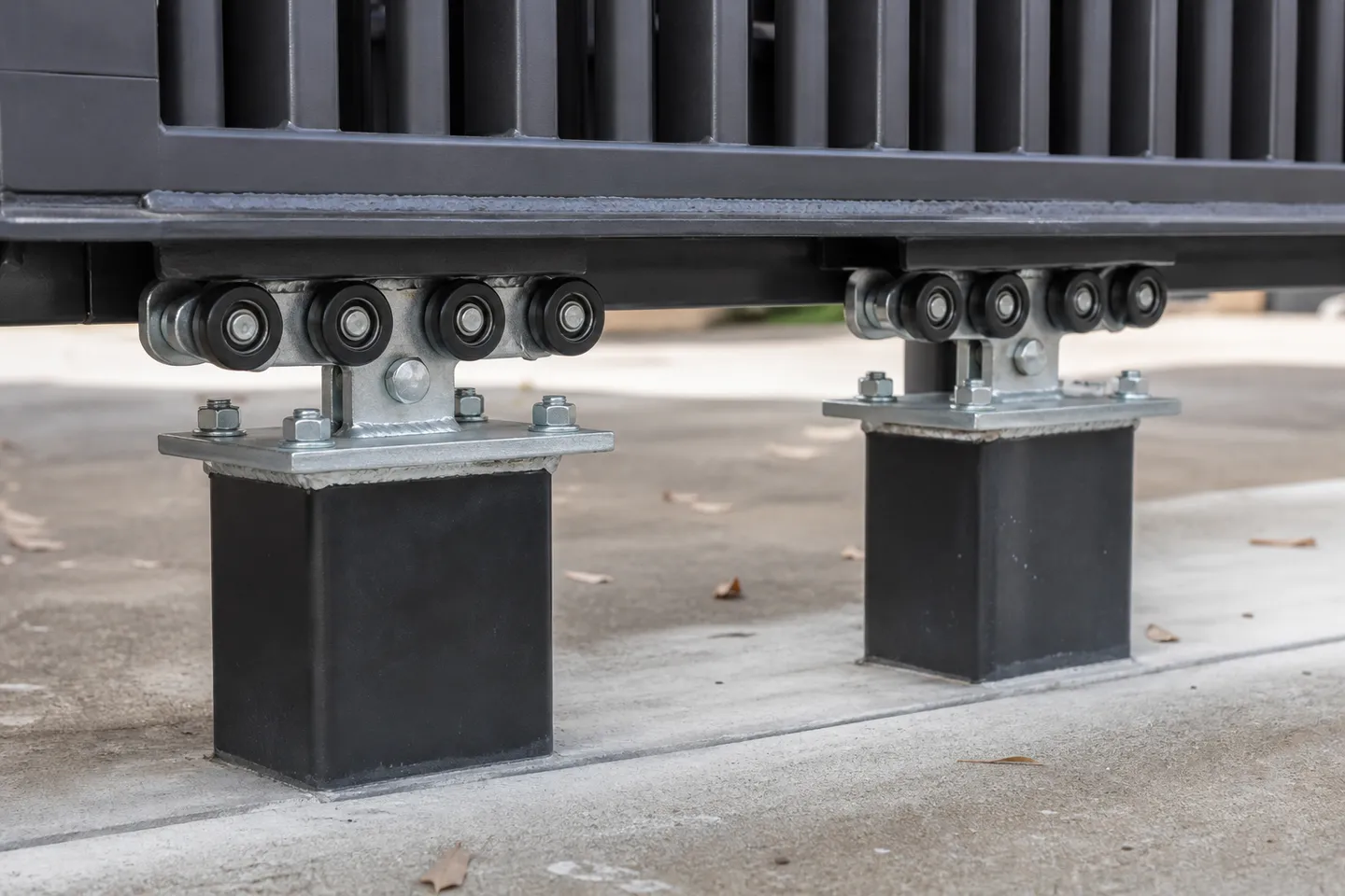 Close-up of two cantilever roller carriages bolted to a square black post, running inside a black aluminium channel under the bottom rail of a sliding gate