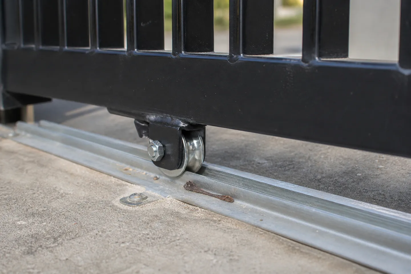Close-up of a grooved sliding gate wheel on a bracket under a black aluminium bottom rail, sitting on a galvanised V-track fixed to a concrete driveway