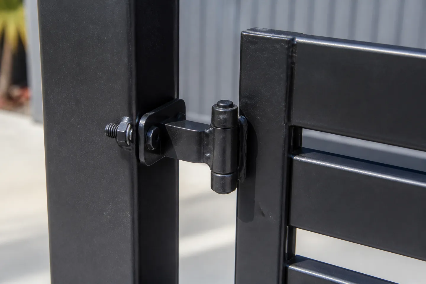 A black adjustable bolt-on gate hinge on a powder-coated aluminium gate, its threaded shank passing through the square post with a nut on each side