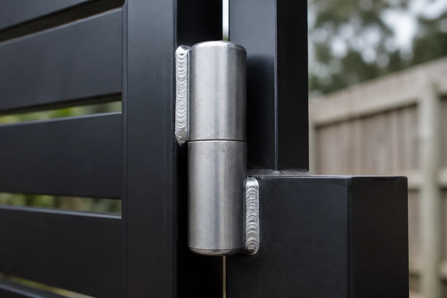 A weld-on bullet hinge joining a black powder-coated gate stile to a square post, with a bright rippled weld bead on each half