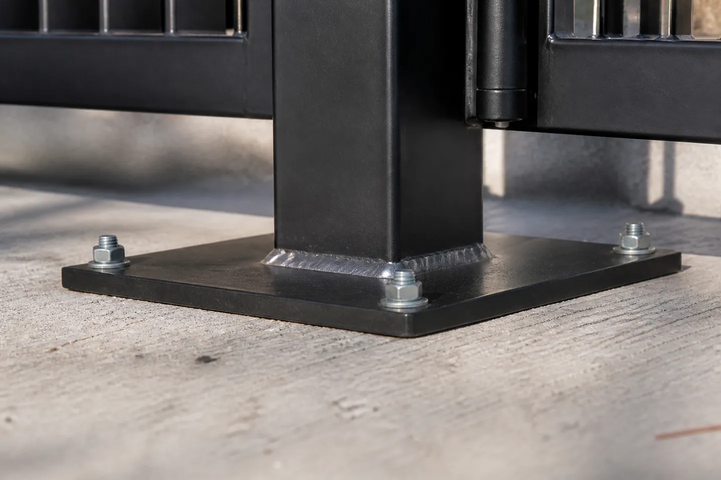 The base of a black square aluminium gate post welded to a square footplate, fixed to a concrete driveway slab with four anchor bolts