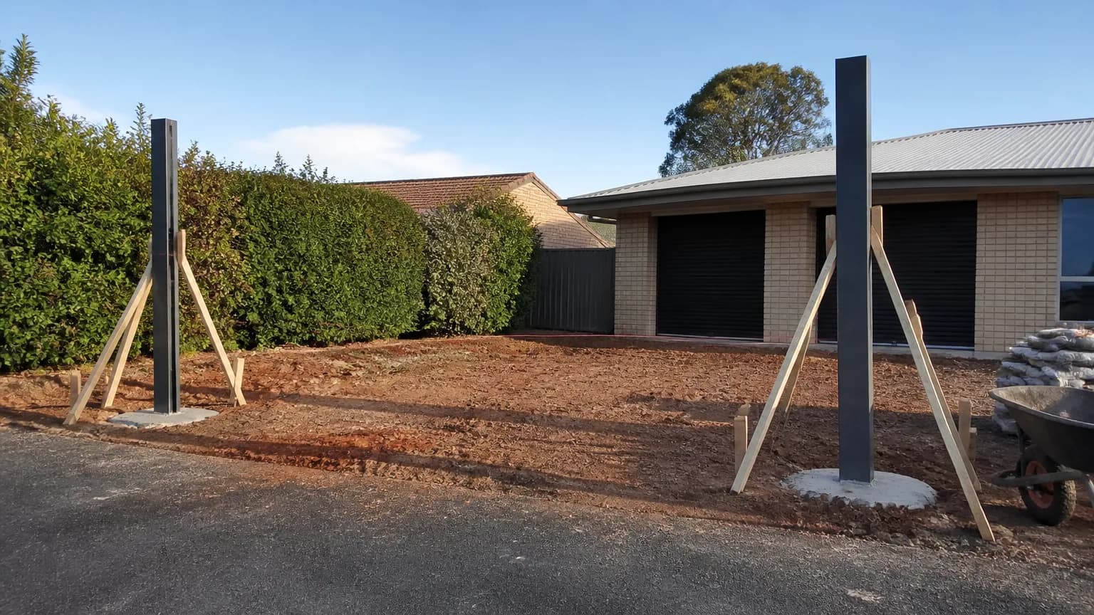 Two black square aluminium gate posts set in fresh round concrete footings across a driveway opening, propped plumb with timber braces