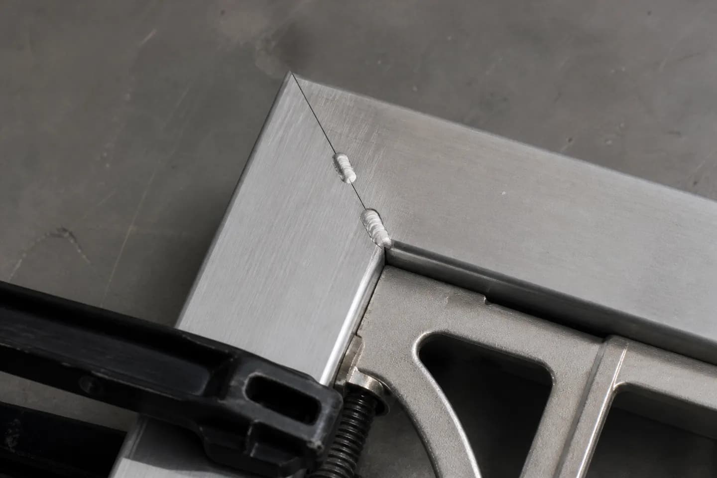 Close-up of a mitred aluminium square tube corner held with a framing square and a clamp, with two small bright tack welds across the joint