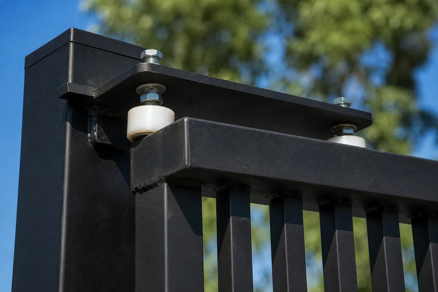 Two nylon guide rollers on a plate bracket at the top of a square black post, one each side of the top rail of a black aluminium sliding gate