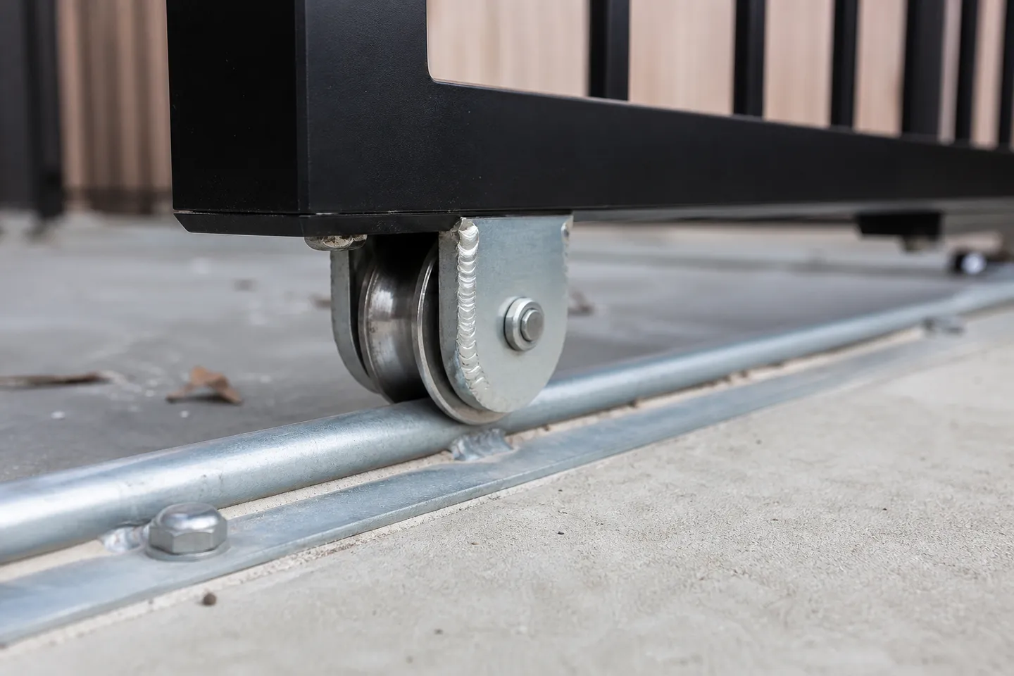 A grooved metal gate wheel in a welded bracket under a black aluminium bottom rail, sitting on a round bar track bolted to a concrete strip