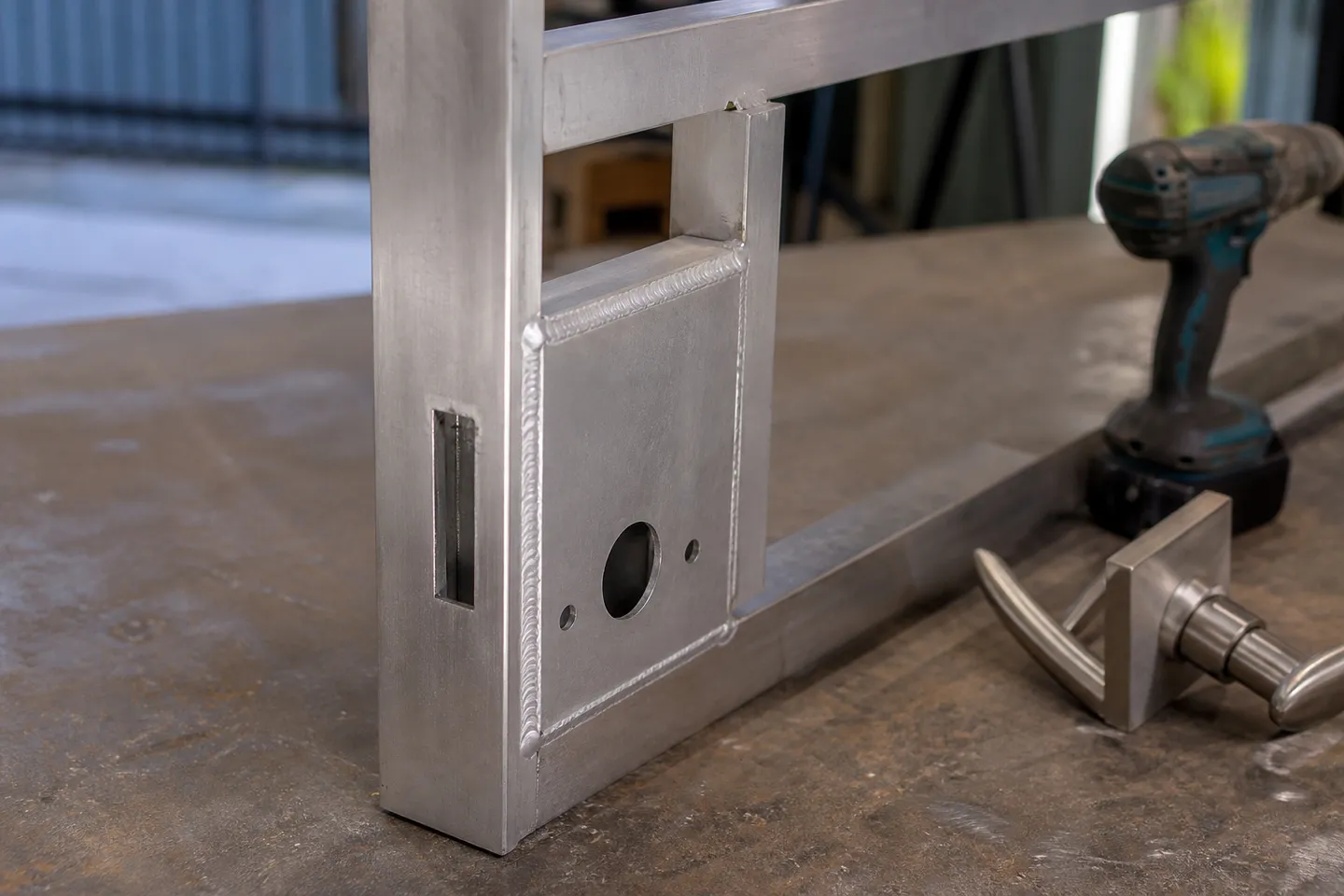 A bare aluminium lock box TIG welded to the latch stile of a gate frame on a workshop bench, with round cylinder and spindle holes drilled through its face
