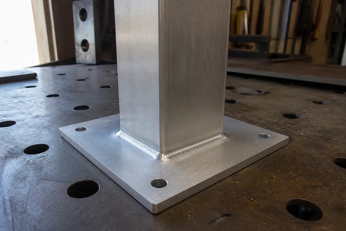 A square aluminium post welded to a flat aluminium base plate with a bright TIG fillet weld running all the way around the post