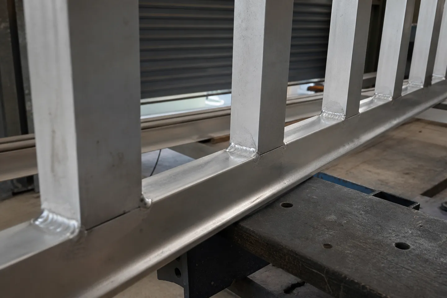 A row of short, evenly spaced TIG stitch welds joining a flat aluminium batten to a square aluminium tube frame rail