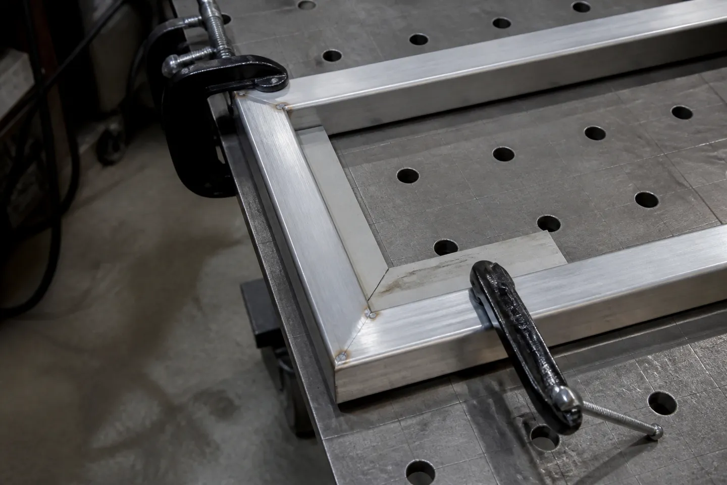 An aluminium square tube gate frame corner held flat to a heavy fabrication table with two F-clamps and a framing square