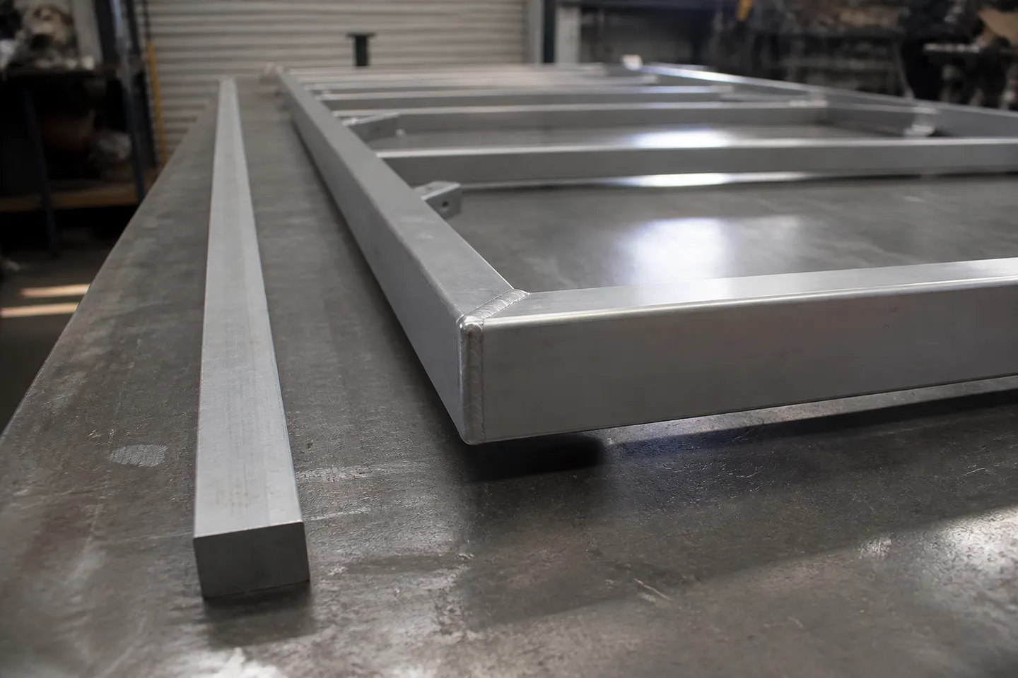 A welded aluminium gate frame lying on a flat fabrication table with one corner lifted clear of the table top, showing a twist