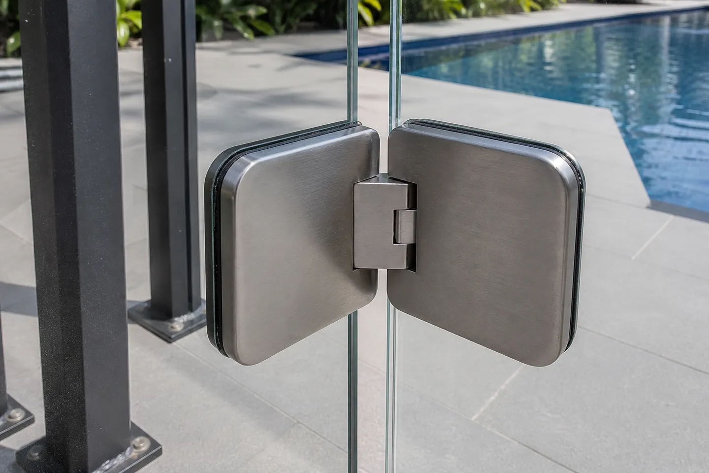 A brushed stainless soft-close hinge clamped between two frameless glass pool fence panels, a pool deck blurred behind