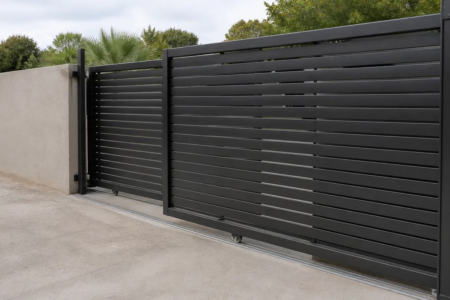 A two-leaf black aluminium slatted telescopic sliding gate fully open, its two leaves stacked side by side on two parallel ground tracks beside a driveway opening